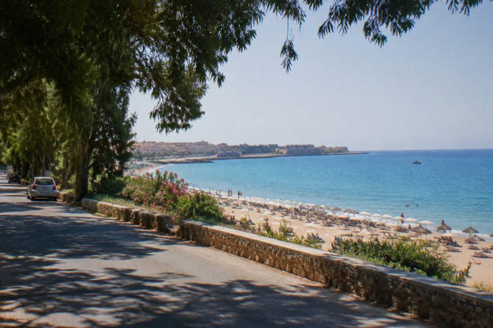 A coastal road above a long sandy beach with umbrellas and calm blue sea beyond