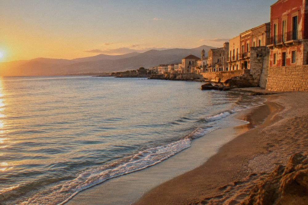 An empty beach on the north-west coast of Crete in late afternoon light, with calm shallow water and hills in the distance