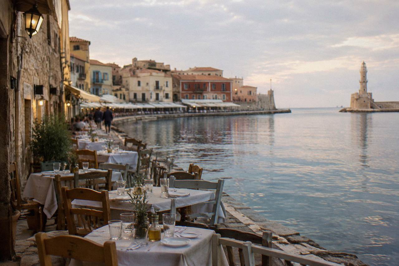 Tables set with glasses outside tavernas along the stone buildings of Chania's old harbour at golden hour