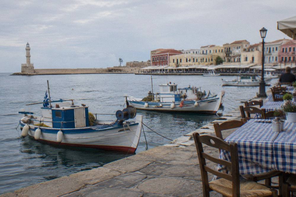 A stone harbour front with wooden fishing boats moored beside cafe tables in the late afternoon