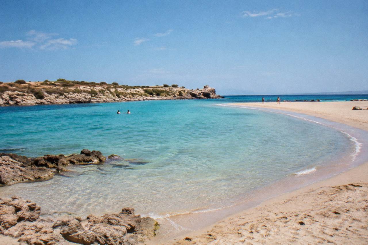 A turquoise lagoon with shallow water curving between a sandy spit and rocky headlands under a clear sky