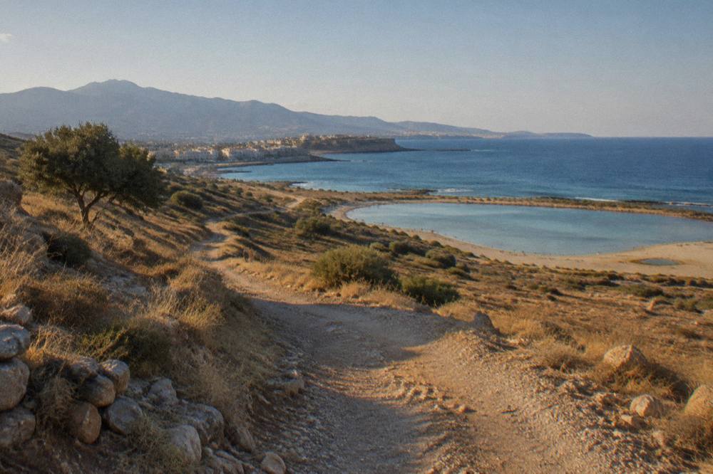 A rough dirt track winding across dry golden hills toward a distant lagoon and the sea