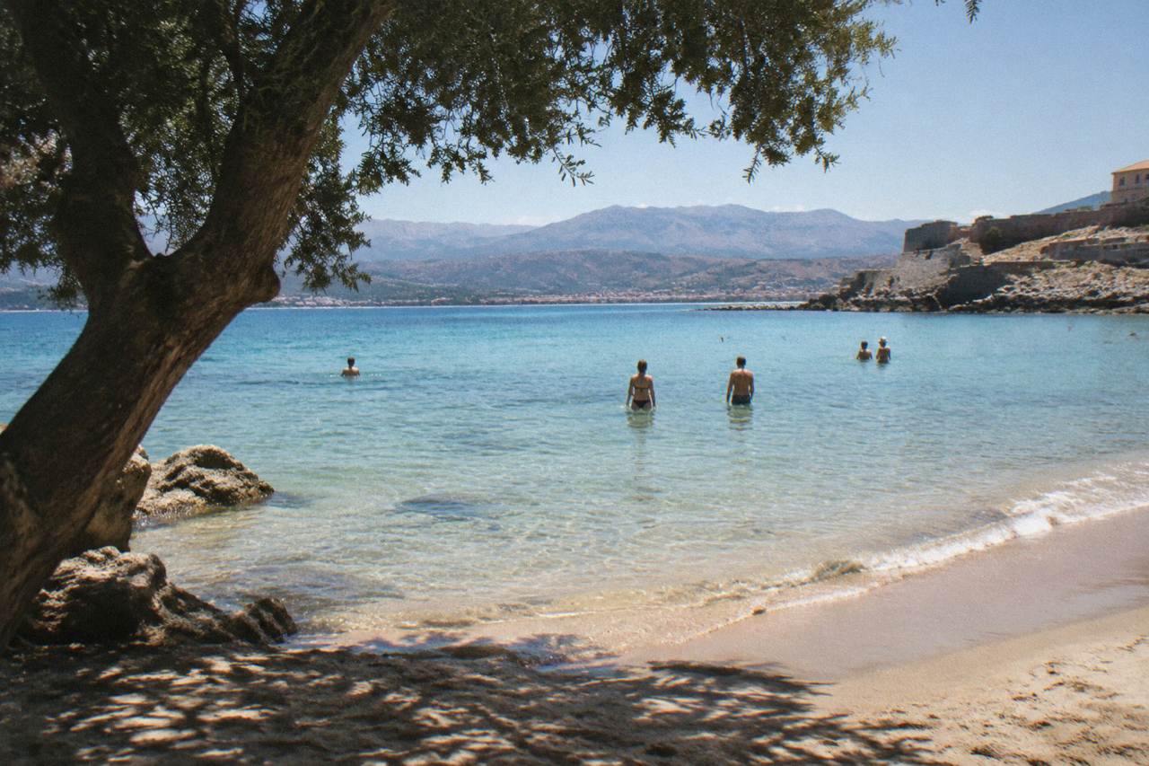 Swimmers standing in clear shallow water at a sandy beach near Chania with hills in the background