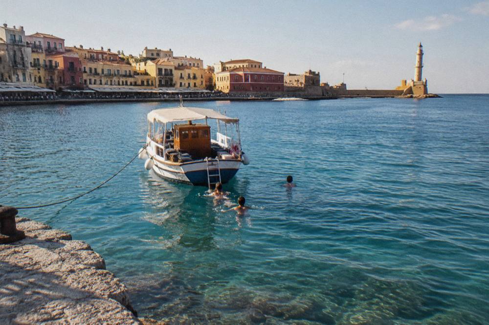 A small boat anchored in a turquoise bay near Chania with people swimming from the stern