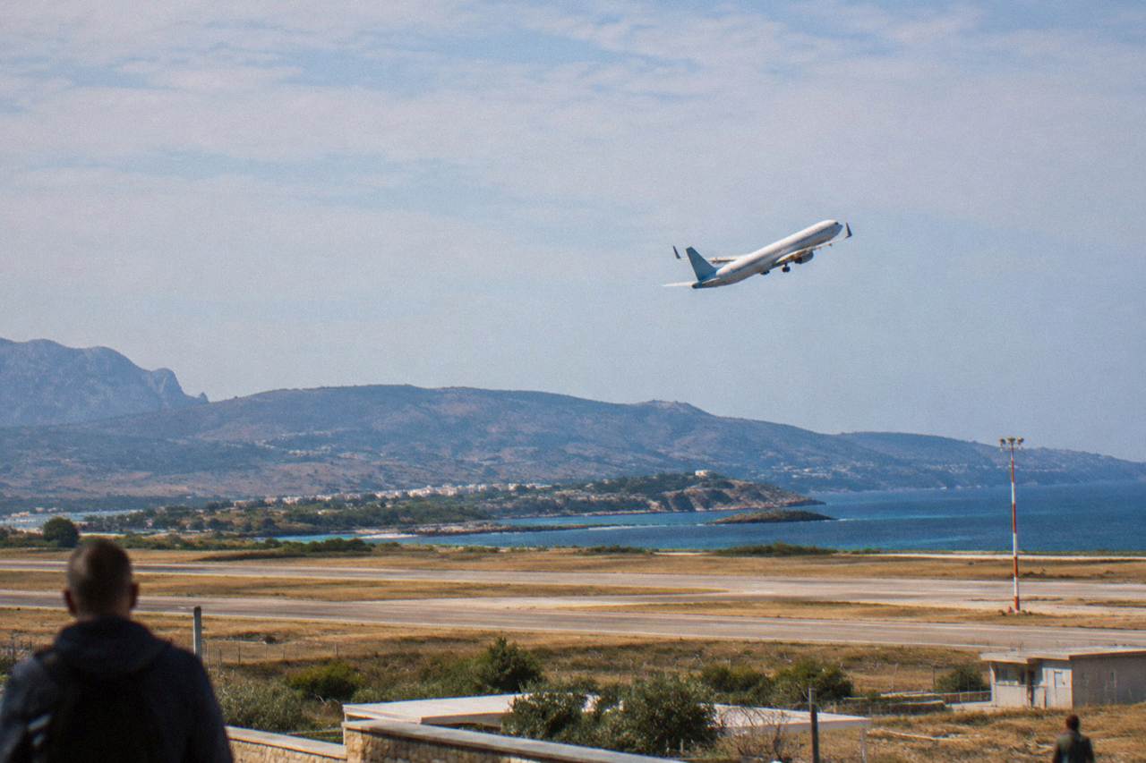 A passenger aircraft climbing over a coastal airport with dry hills and the sea in the background