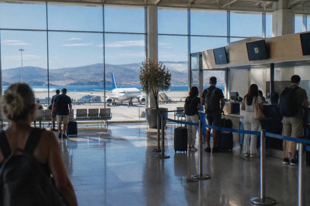 The departure hall of a small Mediterranean airport with travellers at check-in desks and aircraft visible through the glass