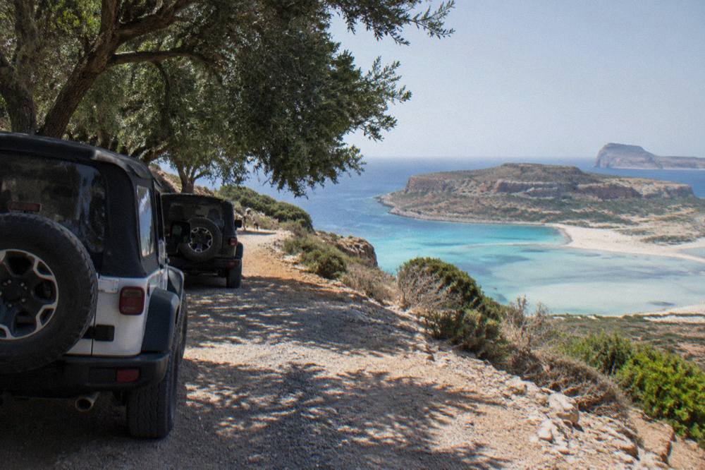 Parked jeeps on a rough gravel track above a coastal lagoon in western Crete