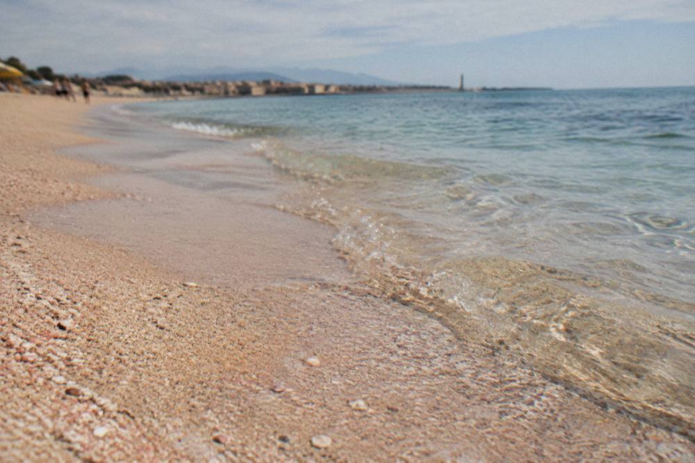 A close view of wet shoreline sand showing pale pink grains mixed with white sand beside clear shallow water