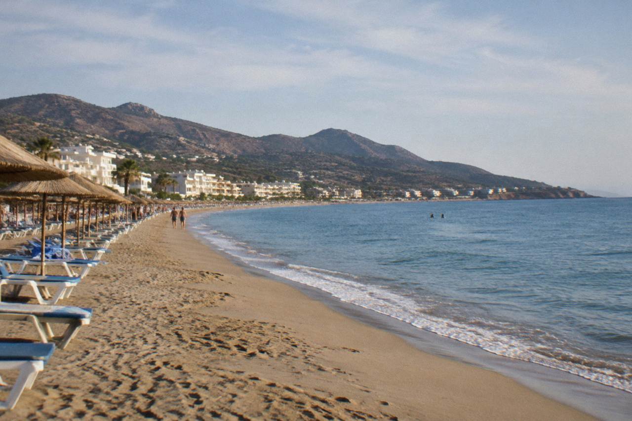 A long sandy beach with rows of sunbeds and calm blue water, hills rising behind a line of small hotels