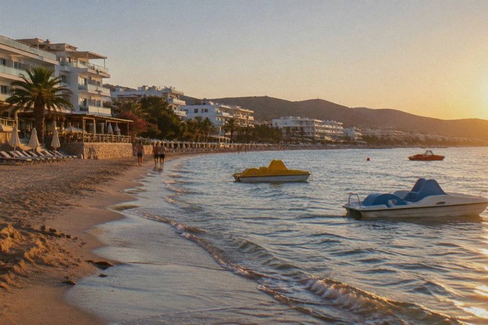 White apartment blocks and hotel terraces above a sandy shoreline with pedal boats in shallow water