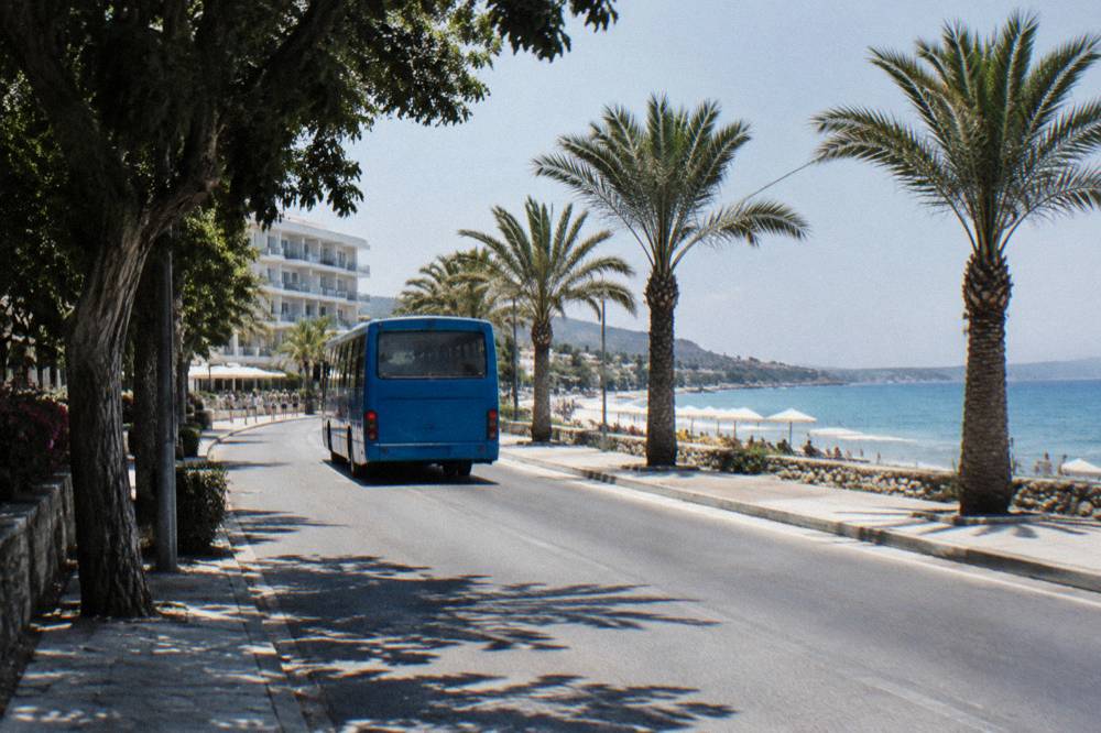 A blue local bus on a coastal road with palm trees and hotel, sea to the right