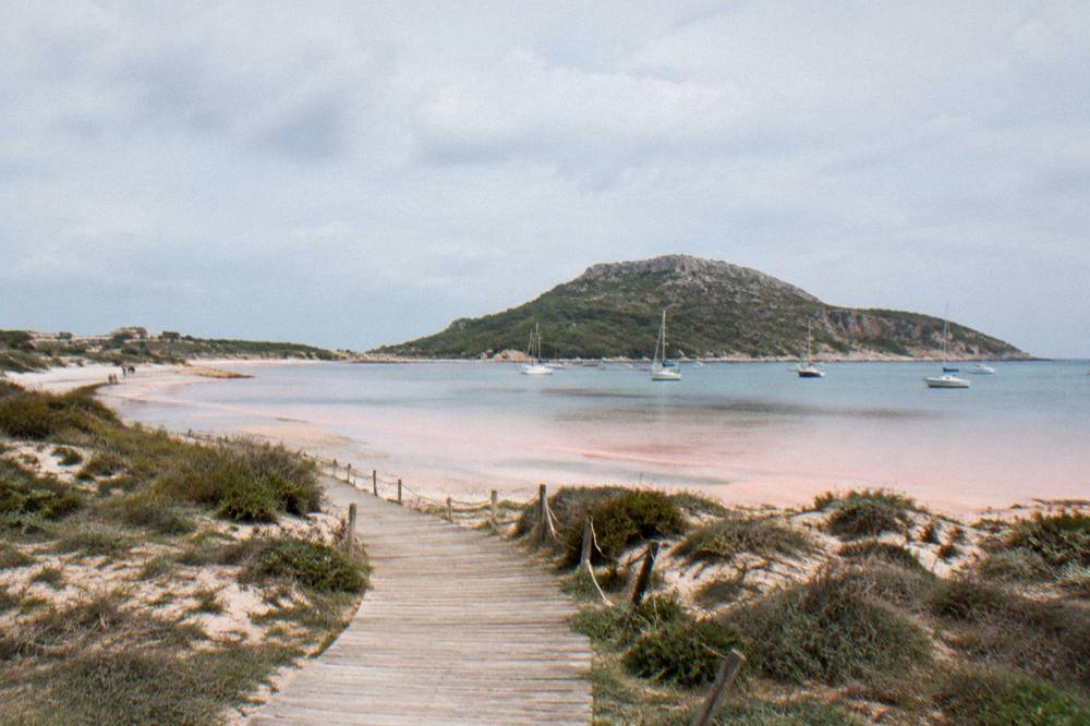 A pink-white lagoon beach with a wooden walkway and an island hill behind, boats at anchor