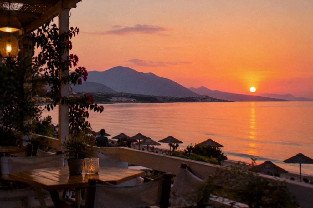 Sunset over the sea seen from a beachfront terrace, mountains turning purple behind a darkening shoreline