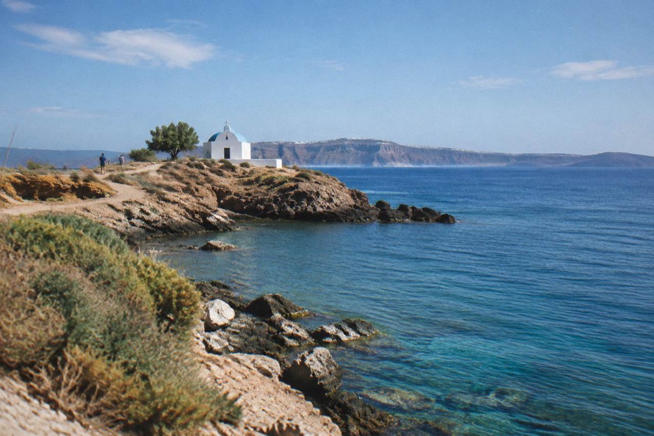 A rocky peninsula coastline with calm blue water and a small chapel on the headland