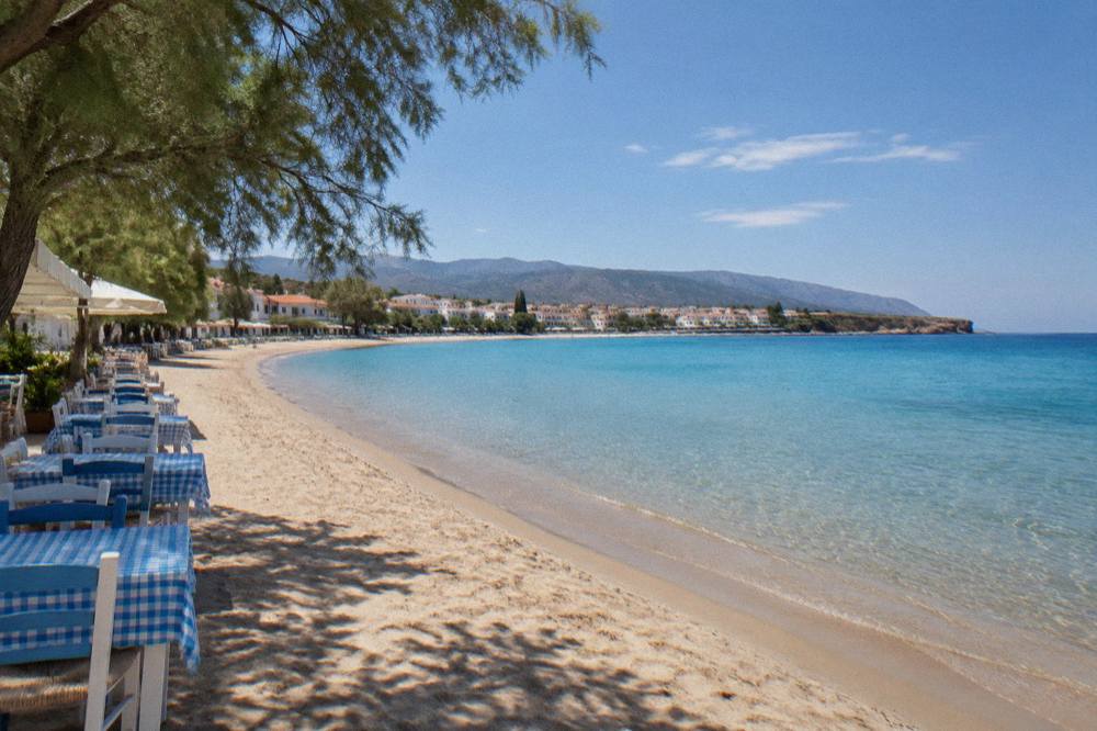An empty sandy beach in a curved bay with calm shallow water and a line of taverna chairs