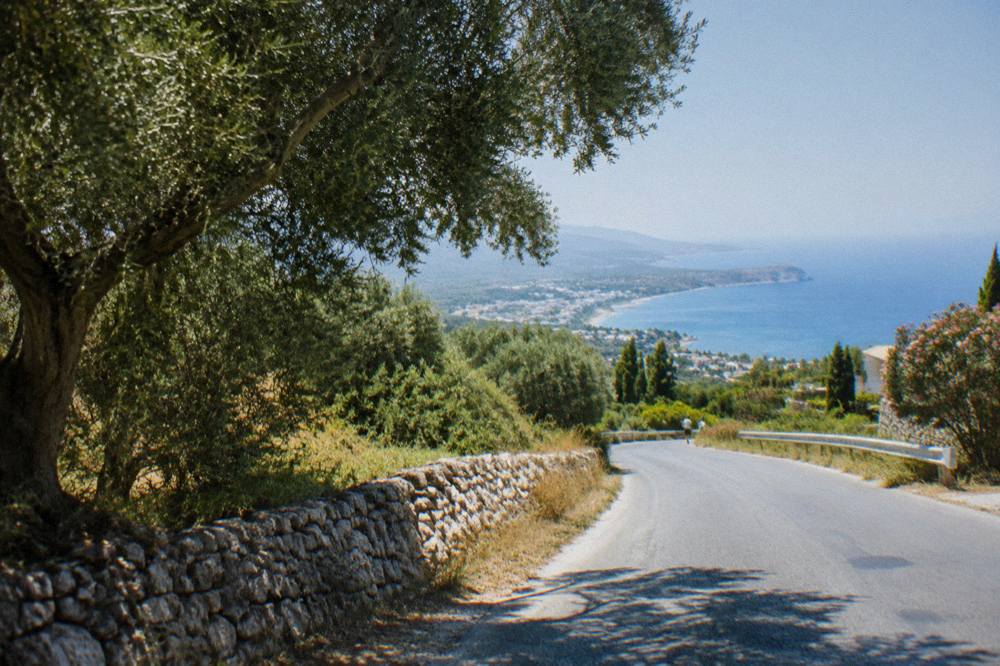 A winding road through green foothills with a glimpse of the sea far below