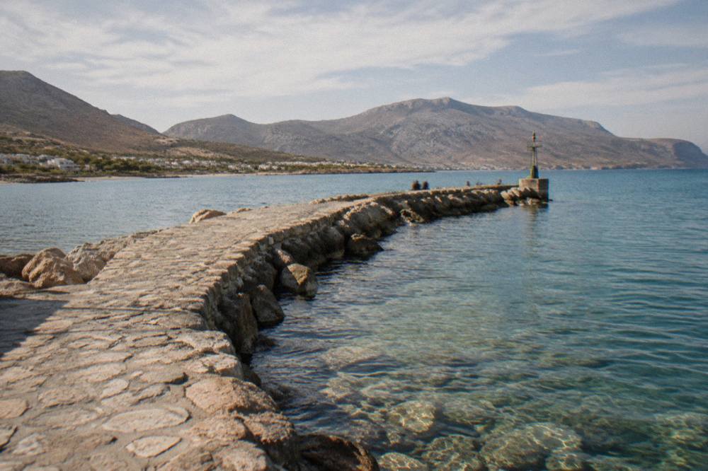 A stone jetty curving into clear shallow water with hills in the background under a bright sky