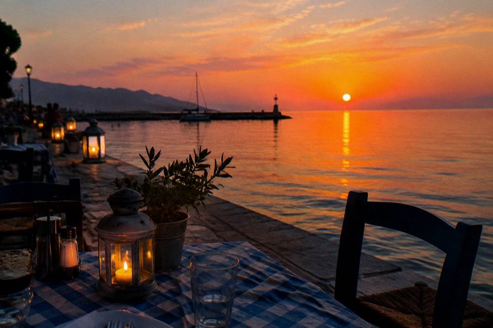 A taverna table set beside the water at dusk with lanterns glowing along a waterfront promenade