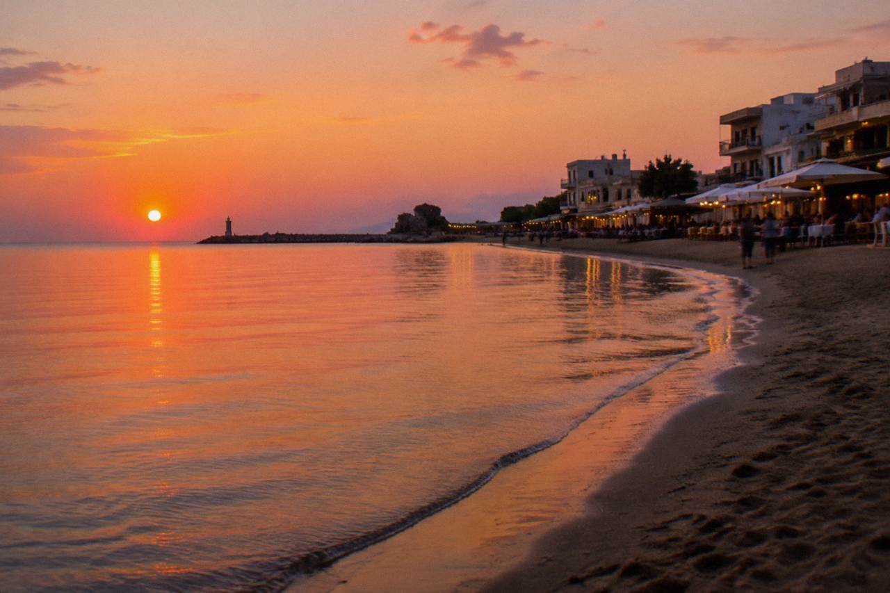 A sandy city beach with calm shallow water and a row of tavernas along the shore in the evening