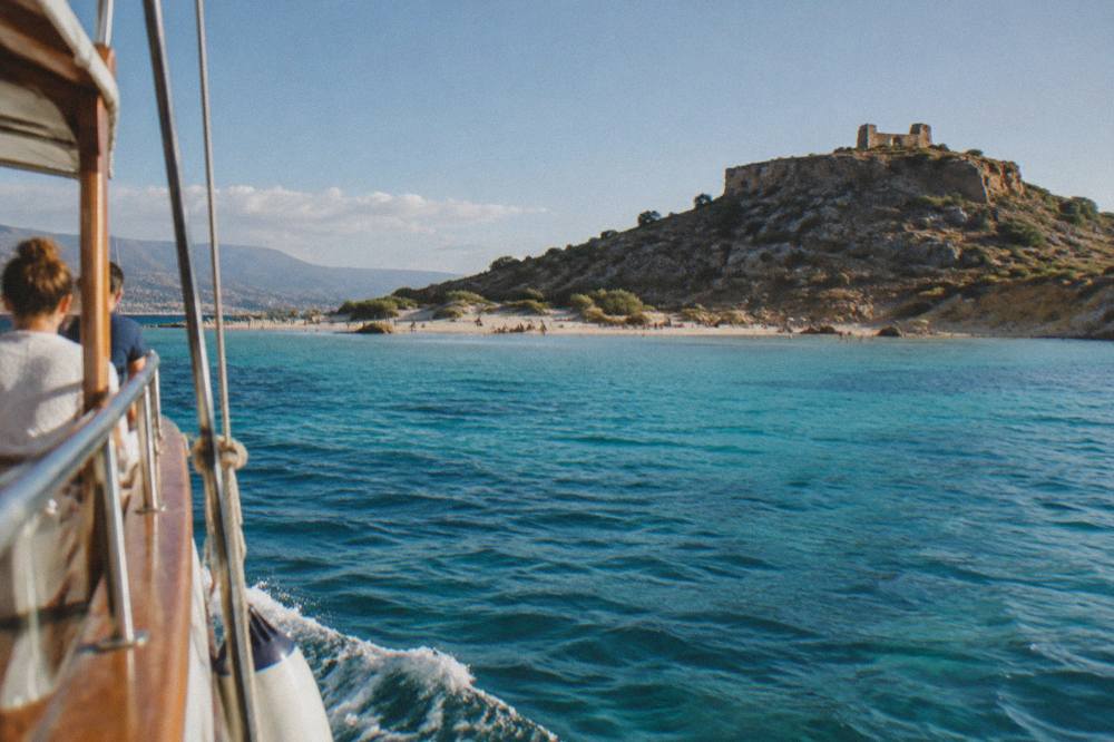 A boat at sea approaching a lagoon beach with turquoise water and a hilltop ruin