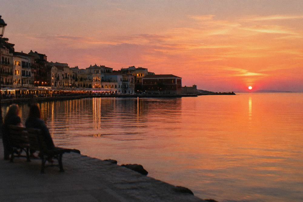 An evening sky over a coastal town with lit windows and the sea calm in front