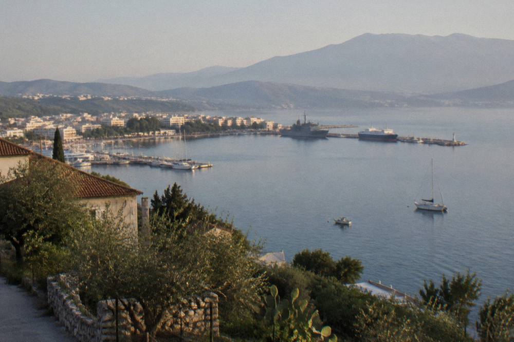 A small port town seen from a hillside, with moored vessels and calm grey-blue water below