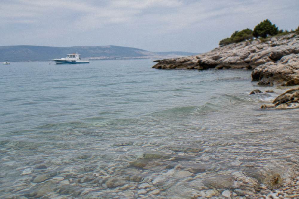 Clear shallow water over pebbles, with a rocky headland and a parked boat in the distance