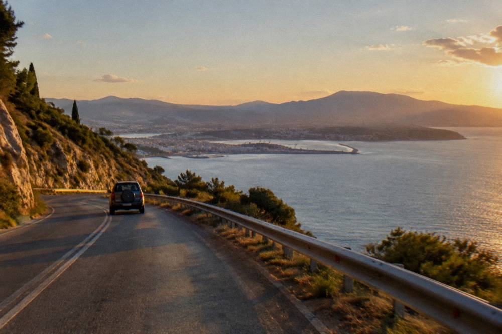 A coast road curving around a hillside above a wide bay, with a vehicle ahead and open water beyond