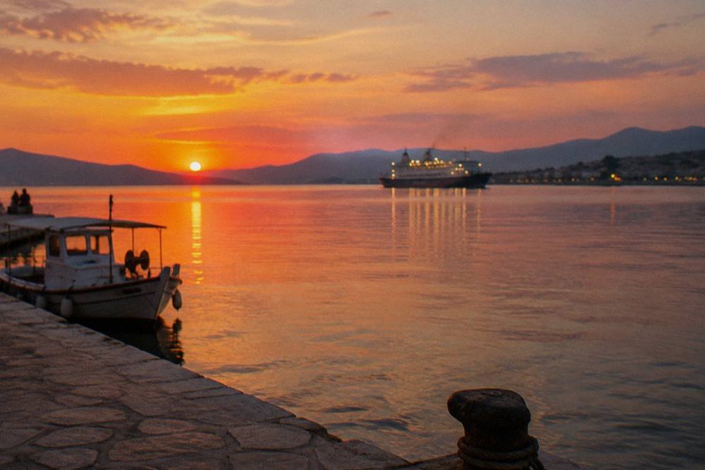 A stone quay at dusk with warm light on the water and a ferry's lights out in the bay