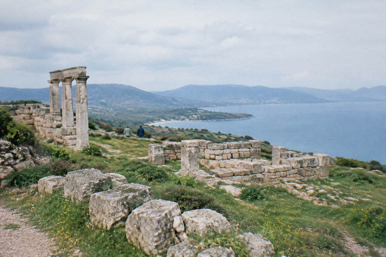 Stone ruins of an ancient site standing on a green hillside above a wide blue bay