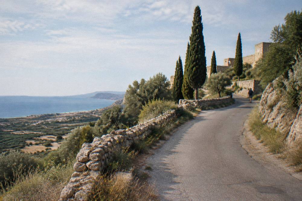 A narrow country road curving up a hillside toward stone walls and cypress trees