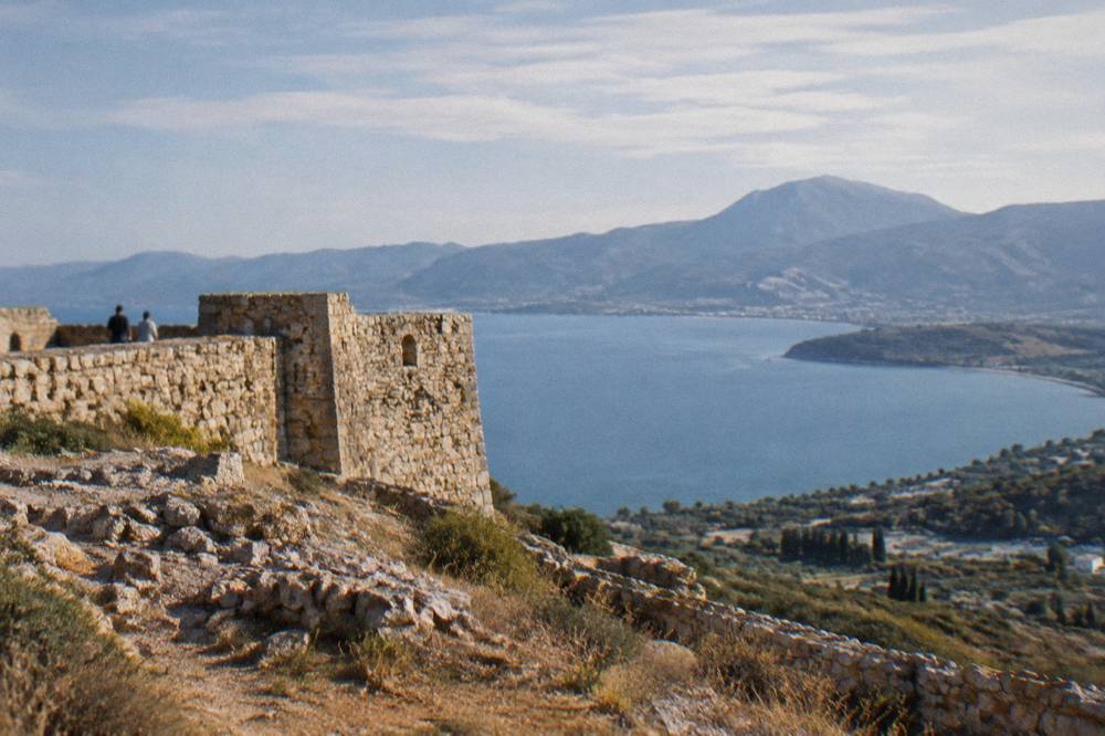 An old stone fortress wall on a hilltop with a wide bay and distant mountains behind it