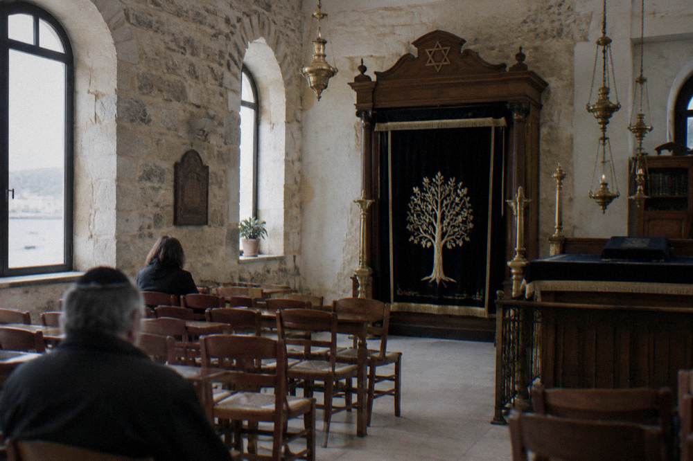 An interior prayer hall with stone walls, a curtained wooden ark, brass lamps and rows of simple chairs