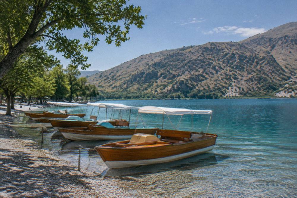 Wooden pedal boats moored in shallow water beside a gravel beach under hillside trees