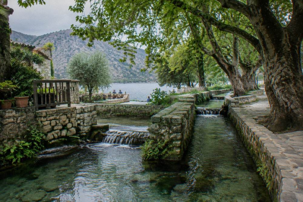 Clear spring water running through stone channels beneath large shade trees in a hill village