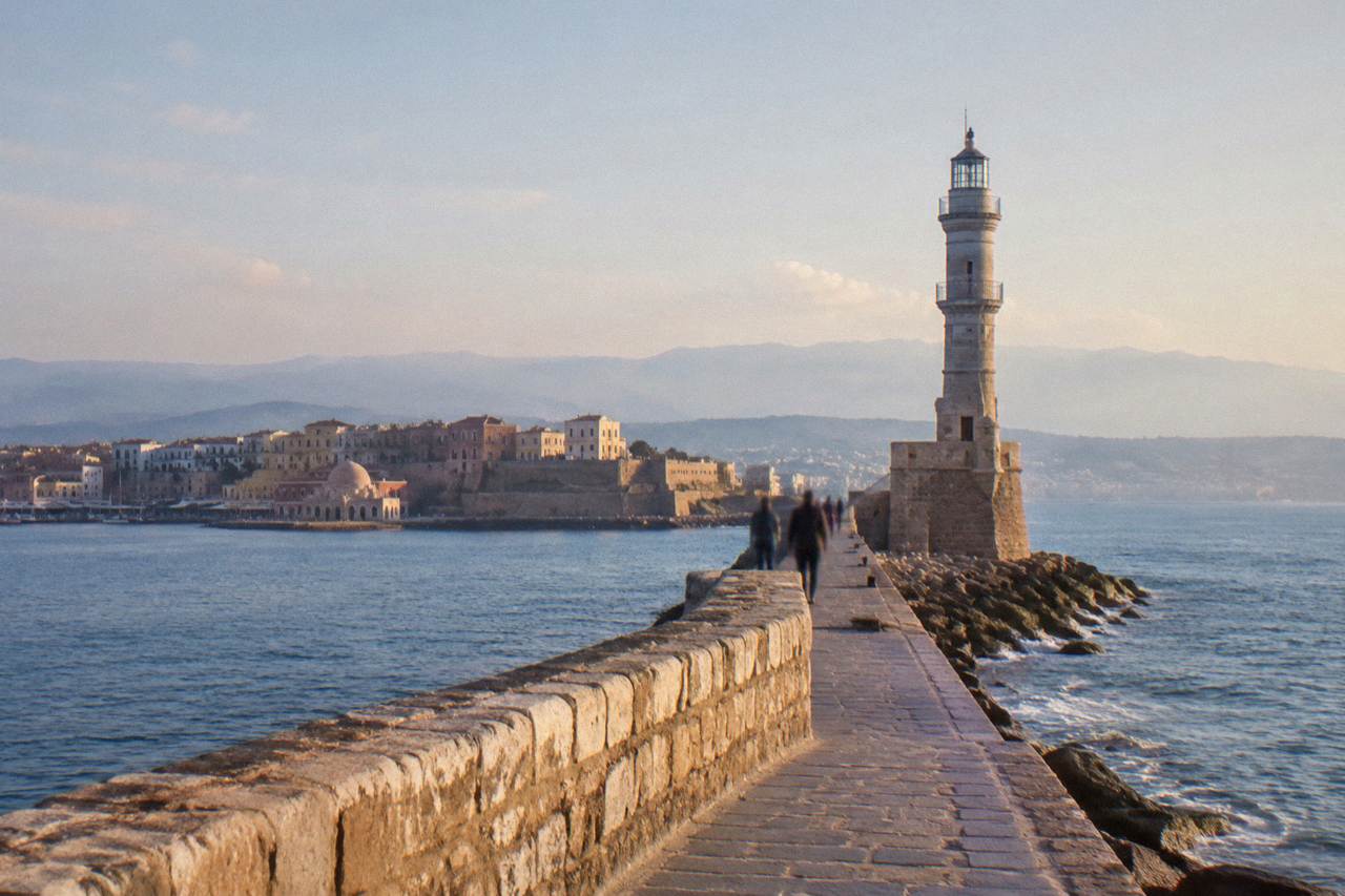 A stone lighthouse standing at the end of a long harbour mole, with the sea on both sides and a city behind it