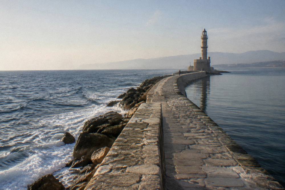 A stone mole path curving out over the sea toward a lighthouse, with waves on the outer side and calm water inside