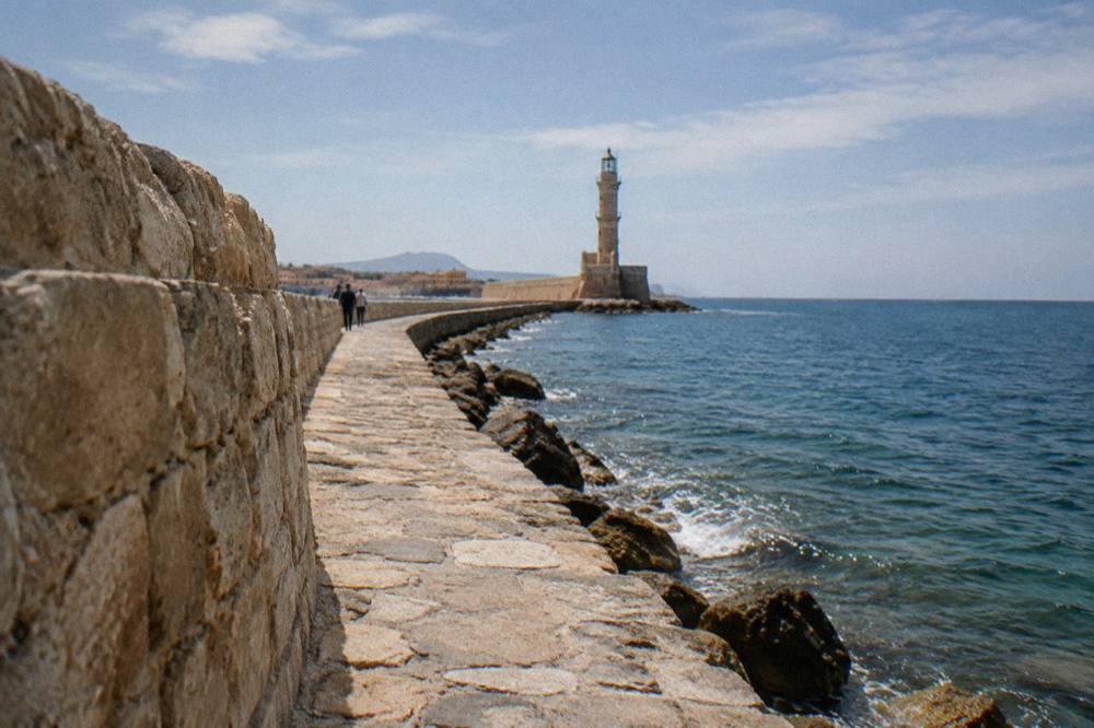 A rough stone path along a sea wall curving toward a distant tower under a clear midday sky