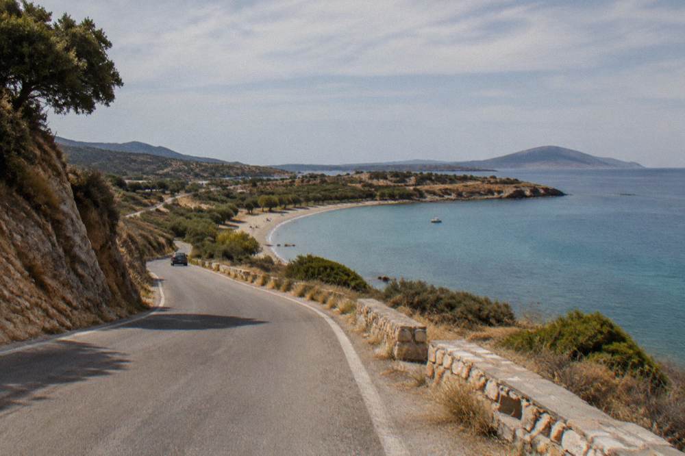 A coastal road curving along a dry hillside toward a small sandy bay