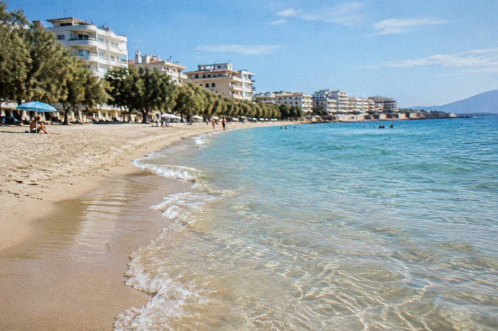 Shallow clear water over pale sand with apartment buildings and tamarisk trees behind the beach