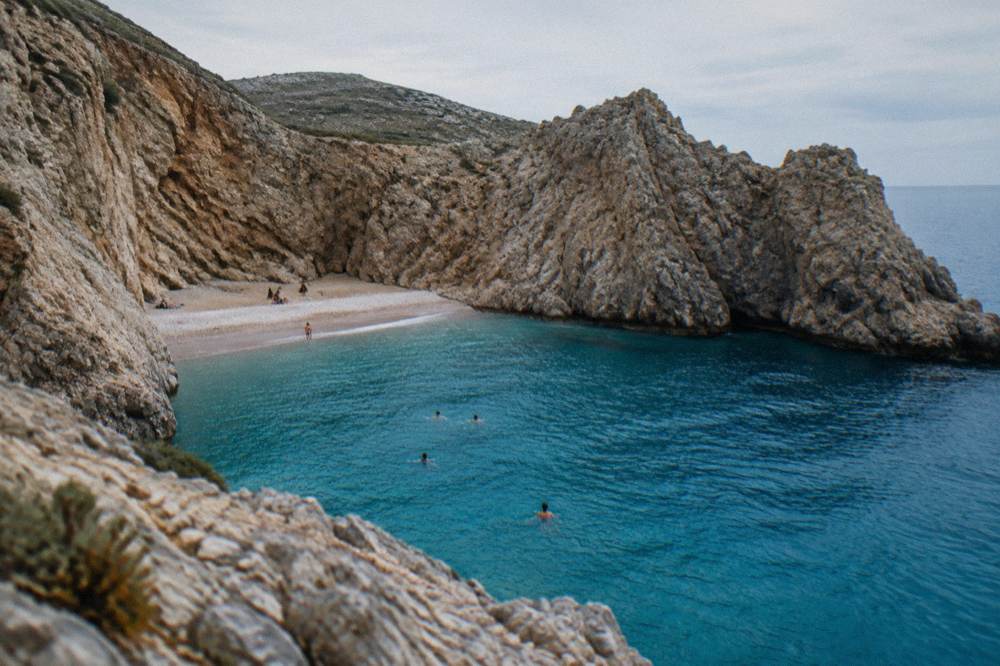 Swimmers in deep blue water beside a narrow strip of sand between cliffs