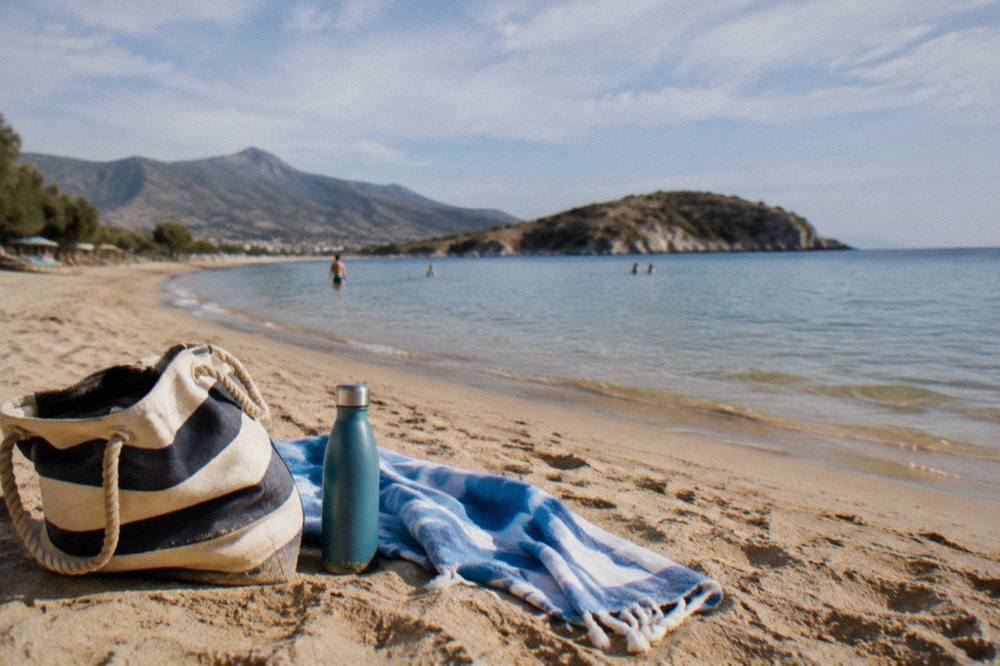 A beach bag, towel and water bottle on sand beside calm shallows