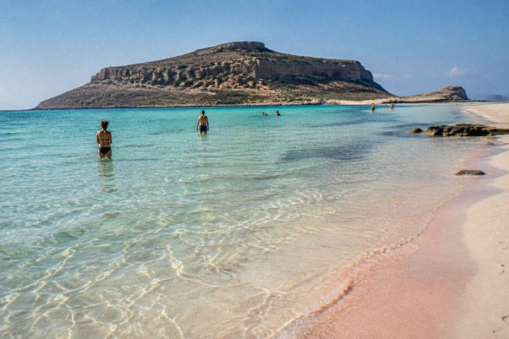 People wading in pale turquoise shallows beside a curve of white and pink sand
