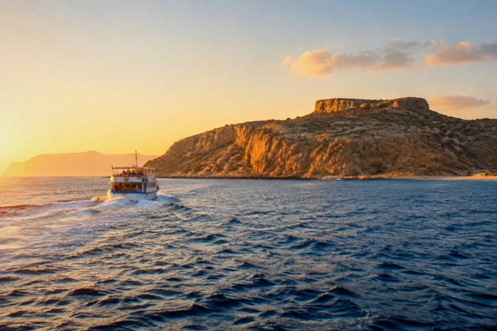 An excursion boat crossing open blue water toward a peninsula at golden hour