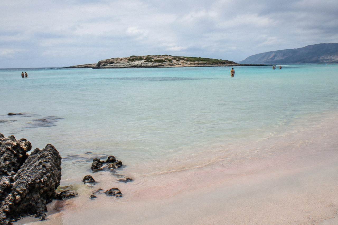 A wide lagoon beach with shallow turquoise water and pale pink sand stretching to a small island