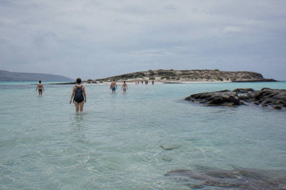 People wading through knee-deep clear water on a sandbar between a beach and a small island