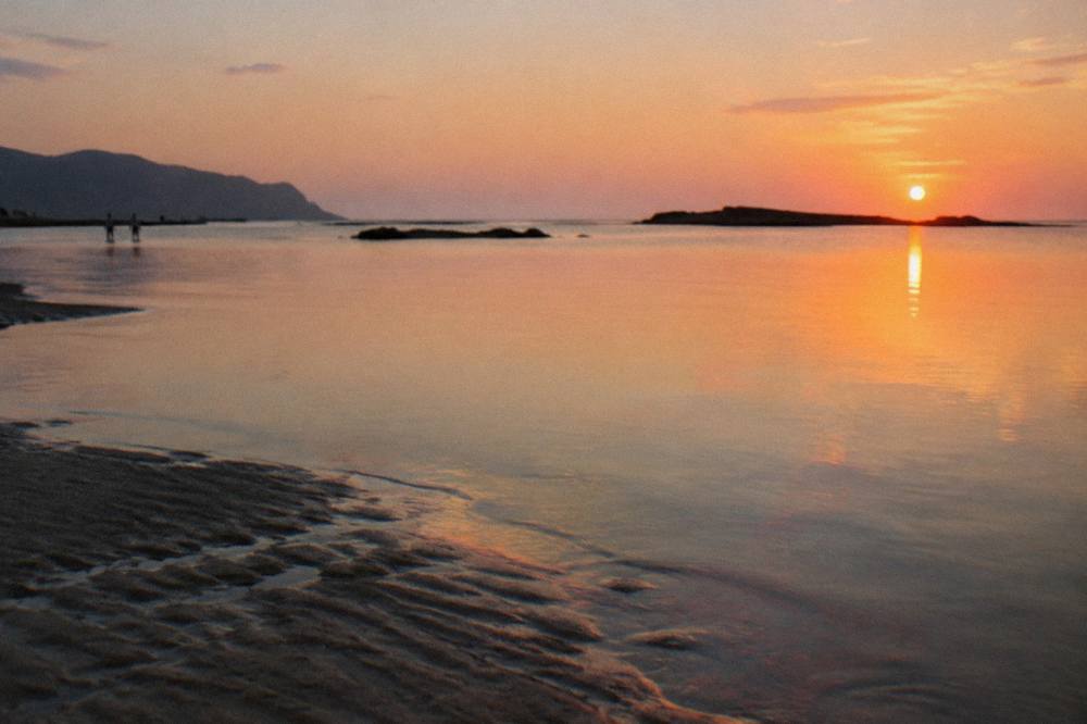 A calm lagoon at sunset with glassy shallows reflecting a pale sky above darkening sand