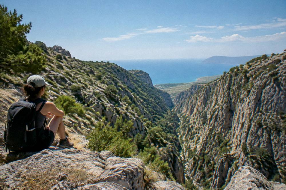 A walker resting on a rock ledge looking out over a deep gorge toward distant sea