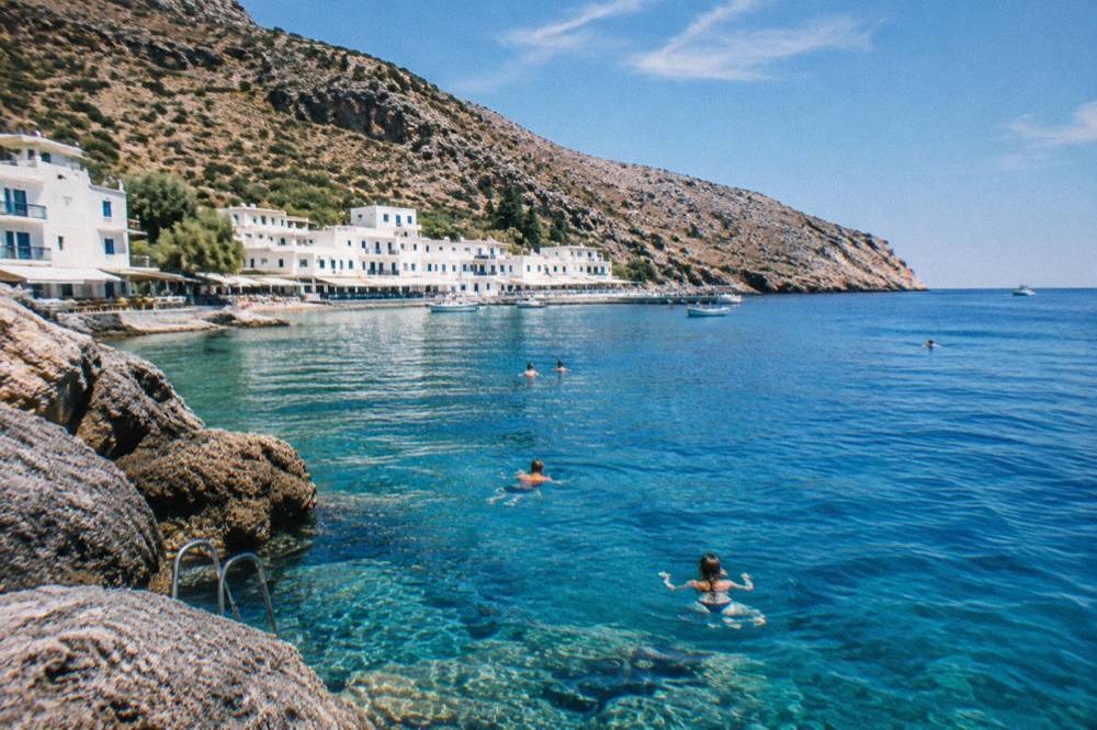 Swimmers in clear deep blue water beside rocks near a whitewashed village