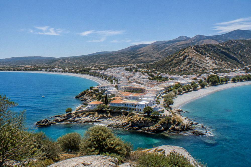 A peninsula town seen from above with beaches on both sides and hills rising behind